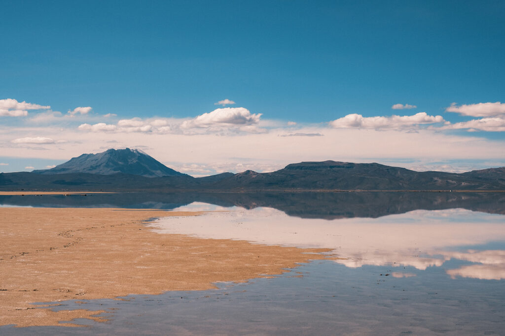 Wide view of the Salinas y Aguada Blanca National Reserve near Arequipa, Peru, with still reflective water, open high-altitude plains, and mountains beneath a bright blue sky.