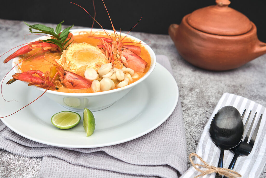 Traditional chupe de camarones from Arequipa, Peru, served in a white bowl with river shrimp, egg, corn, lime, and a clay pot on the side.