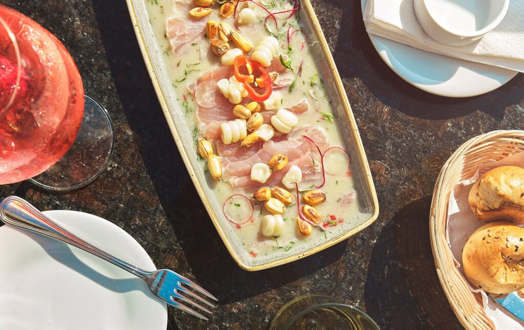 Tiradito de pescado served in a creamy sauce with Peruvian corn, toasted cancha, sliced chili, and fresh herbs on a restaurant table in Lima.